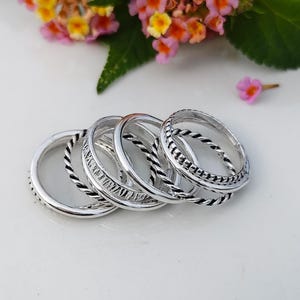 Könnte beinhalten: Ein Set aus fünf silberfarbenen Ringen, die in einem überlappenden Stapel angeordnet sind. Die Kollektion umfasst Ringe mit glatten Oberflächen, gedrehten seilartigen Texturen und einen Ring mit einem detaillierten Ätzmuster am Außenrand.