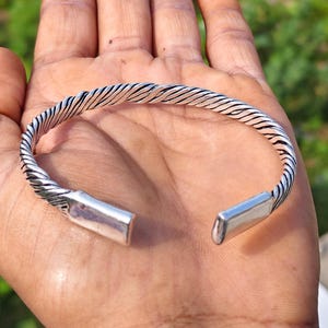 Könnte beinhalten: Ein silberfarbenes Armband in Manschettenform mit einer gedrehten, seilartigen Textur entlang des Reifs und glatten, flachen, rechteckigen Endstücken an beiden Öffnungen. Das Metall hat ein poliertes Finish mit dunkler Oxidation in den Rillen.