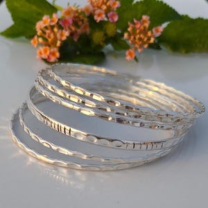 Puede incluir: Una pila de cinco brazaletes de color plateado con una superficie martillada y texturizada. Las pulseras presentan un acabado metálico reflectante y un patrón repetitivo de pequeñas hendiduras a lo largo del borde exterior.