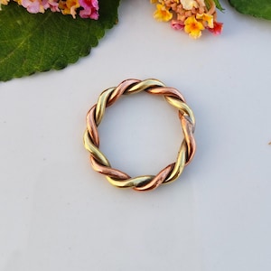 May include: A circular ring constructed from three intertwined metal strands. The design features alternating strands of polished gold-tone metal and warm copper-tone metal, creating a continuous twisted pattern throughout the entire band.