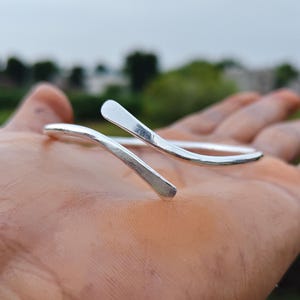 May include: A silver-colored open cuff bracelet with a smooth, polished finish. The bracelet features a bypass design where the two ends overlap slightly, with each end tapering to a flattened, rounded point. The metal has a reflective surface.