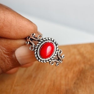 May include: A silver-toned ring featuring an oval-shaped, smooth red stone set in the center. The metal band is decorated with a twisted rope-like border around the stone and intricate, open-work scroll patterns extending along the sides.
