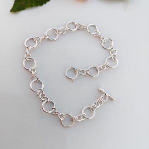 May include: A silver-toned chain bracelet constructed from a series of interconnected circular links of varying sizes. The bracelet features a toggle clasp closure consisting of a small horizontal bar that slides through a larger circular ring.