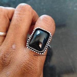 May include: A silver ring featuring a large, rectangular, polished black stone set in a beaded metal frame. The band has a twisted rope texture. The text SilverArtisanitems is visible on the lower portion of the ring setting.