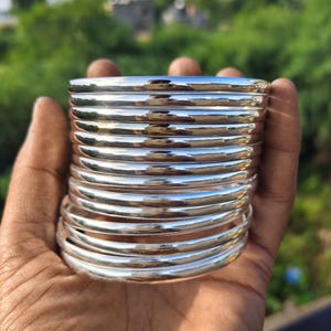 Puede incluir: Una pila de doce brazaletes plateados pulidos con una superficie lisa y redondeada. Cada brazalete presenta un acabado metálico reflectante que capta la luz, creando una apariencia uniforme en todo el conjunto de pulseras circulares.