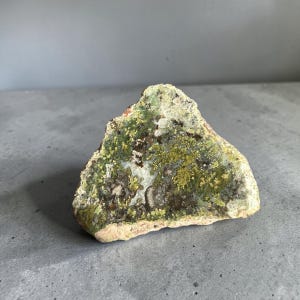 May include: A triangular-shaped rock specimen featuring a textured surface with a mix of moss green, lime green, and dark brown mineral inclusions. The stone has a rough, natural edge and a crystalline appearance with white and translucent patches.