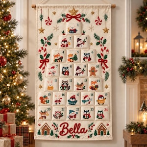 Pode incluir: Um calendário do advento de tecido cor de creme pendurado verticalmente, com vinte e quatro bolsos numerados com personagens natalinos bordados. O nome Bella está costurado em vermelho na parte inferior, rodeado por motivos de casas de gengibre.