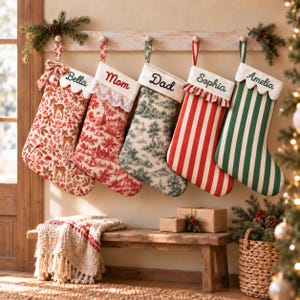 May include: Five personalized Christmas stockings hanging from a wooden rack. The stockings feature various patterns including deer, toile, and stripes in red, green, and white. Each stocking has a white cuff embroidered with names: Bella, Mom, Dad, Sophia, and Amelia.
