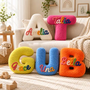 May include: Five plush, textured letter-shaped cushions in cream, pink, yellow, blue, and orange. Each cushion features a colourful, embroidered name: Amelia, Talia, Grace, Una, and Bella. The cushions are arranged on and in front of a sofa.