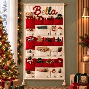 Può includere: Un calendario dell'Avvento in tessuto color crema da appendere, con ventiquattro tasche rosse e crema. Il nome Bella è ricamato in alto in rosso, verde e oro. Ogni tasca è numerata e riempita con piccoli ornamenti natalizi decorativi.