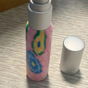 May include: A small cylindrical spray bottle with a pink base featuring a pattern of yellow and blue flowers. The bottle has a silver-colored pump mechanism and a separate matching silver-colored cap resting beside it on the surface.