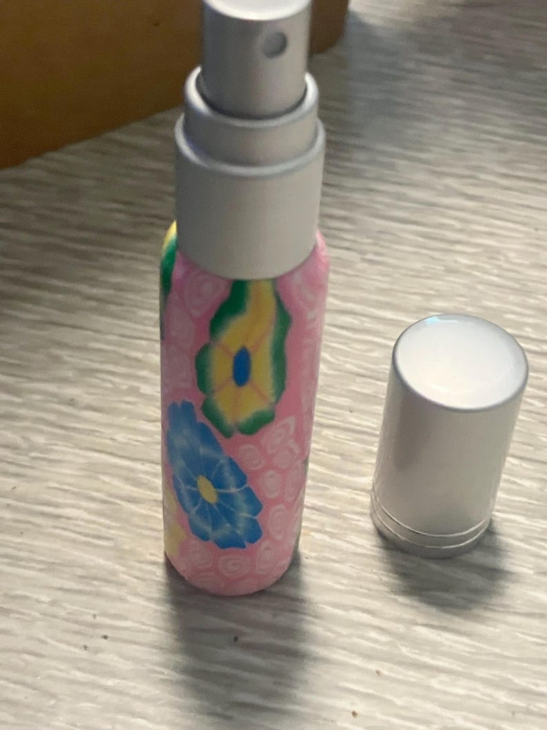 May include: A small cylindrical spray bottle with a pink base featuring a pattern of yellow and blue flowers. The bottle has a silver-colored pump mechanism and a separate matching silver-colored cap resting beside it on the surface.