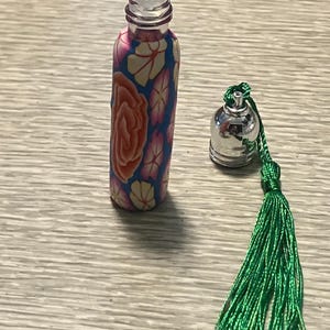 May include: A small cylindrical glass roll-on bottle featuring a colorful floral pattern in shades of orange, pink, and cream against a blue background. Beside the bottle sits a silver-toned metal cap attached to a long, vibrant green tassel.