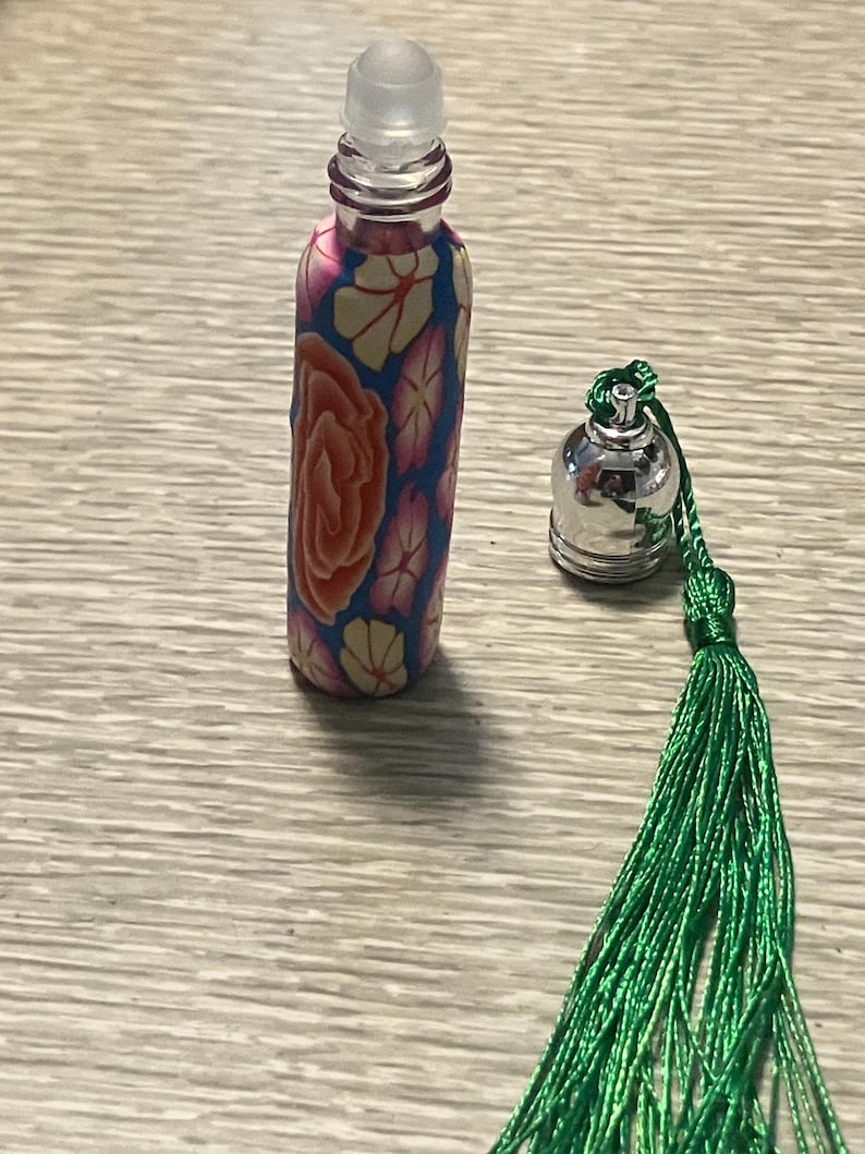 May include: A small cylindrical glass roll-on bottle featuring a colorful floral pattern in shades of orange, pink, and cream against a blue background. Beside the bottle sits a silver-toned metal cap attached to a long, vibrant green tassel.