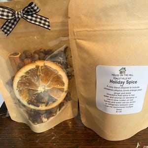 May include: Two brown paper pouches containing Holiday Spice. The left pouch has a clear window showing dried orange slices, cinnamon sticks, and cloves, topped with a black and white gingham bow. The right pouch features a white label with text.