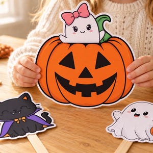 Può includere: Tre decorazioni in carta fustellata raffiguranti una zucca arancione brillante con un fantasma bianco che indossa un fiocco rosa, un gatto nero con un mantello viola e un fantasma bianco che tiene un secchiello a forma di zucca, su bastoncini.