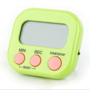 May include: A lime green digital kitchen timer with a rectangular LCD screen and three round pink buttons. The buttons are labeled MIN, SEC, and START/STOP, with the word RESET printed below the first two buttons in black text.