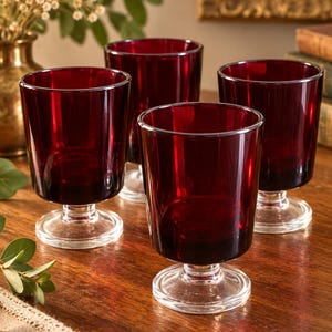 May include: A set of four deep red glass tumblers, each featuring a smooth, cylindrical bowl shape that tapers slightly toward the base. The glasses are supported by a short, clear glass pedestal stem and a circular, flat clear glass foot.