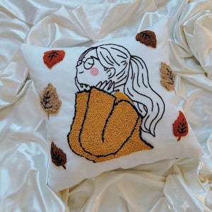 May include: A square white throw pillow featuring a punch needle embroidery design of a person with a ponytail wearing glasses and a mustard yellow sweater, surrounded by several small, textured, multi-coloured autumn leaves in shades of brown and red.