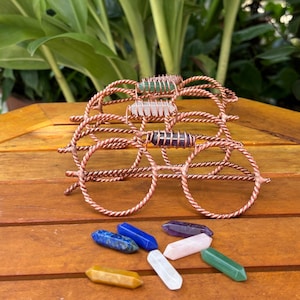 May include: A pair of decorative glasses crafted from twisted copper wire, featuring a horizontal bar across the bridge that holds a small, cylindrical crystal. Below the glasses lie several loose, polished, hexagonal crystal points in various colors.