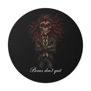 Op de afbeelding: Een ronde zwarte sticker met een skeletfiguur met lang rood haar, gekleed in een donker krijtstreep pak, een wit overhemd, een rode stropdas en een ronde zonnebril met rode glazen. Het skelet heeft gekruiste armen en de tekst Bones don't quit.