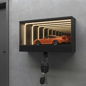 May include: A black rectangular wall-mounted key holder featuring an integrated infinity mirror display with warm LED lighting. An orange sports car model is positioned inside the display, and a metal hook holds a set of keys below the unit.