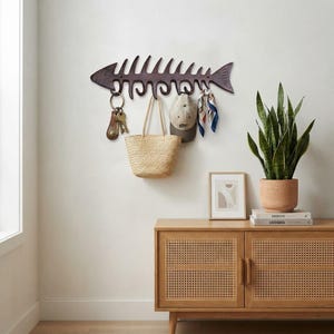 May include: A dark brown, cast iron wall-mounted hook rack shaped like a fish skeleton. The rack features multiple curved hooks holding a set of keys on a ring, a woven straw tote bag, a beige baseball cap, and a patterned silk scarf.
