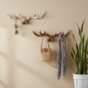Puede incluir: Dos percheros de pared con forma de astas de ciervo. El gancho superior es de color blanco roto y tiene colgadas unas gafas de sol redondas. El gancho inferior es marrón y sostiene un bolso de paja trenzada con asas de cuero y una bufanda gris.