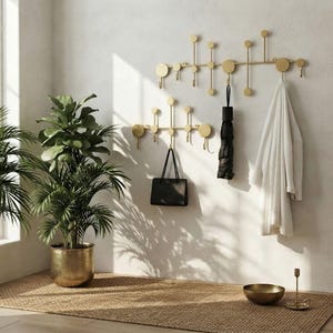 Op de afbeelding: Twee goudkleurige metalen wandkapstokken met een lineair ontwerp, voorzien van meerdere verticale stangen, ronde accenten en haken. Een zwarte handtas, een zwarte paraplu en een witte badjas hangen aan de haken van de wandrekken.