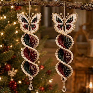 May include: Two hanging crochet ornaments featuring a butterfly top with cream, dark green, and deep red yarn. Below the butterfly, a spiraling, ribbon-like crochet tail in alternating dark green and deep red with gold star embroidery and a clear crystal bead.