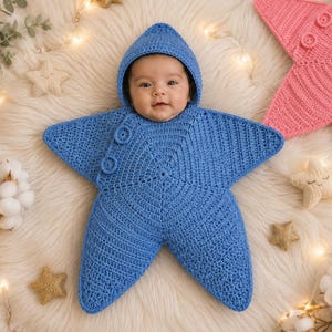 May include: A blue crocheted star-shaped swaddle blanket with an integrated hood. The garment features a textured stitch pattern and two circular buttons on the upper left point. The swaddle is designed to wrap around an infant, leaving the face visible.