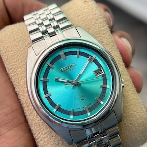 Könnte beinhalten: Eine silberfarbene Seiko Automatikuhr mit einem leuchtend türkisfarbenen Zifferblatt mit Sonnenschliff, schwarzer Minuterie, silbernen Stabindizes, silbernen Zeigern, Datumsanzeige auf der Drei-Uhr-Position und einem Edelstahl-Gliederarmband.