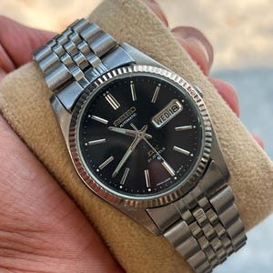 May include: A silver-tone Seiko automatic wristwatch with a black dial, featuring a fluted bezel, a stainless steel link bracelet, and a day-date window at the three o'clock position. The dial displays the text Seiko, Automatic, DX, 21 Jewels.