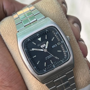 May include: A Seiko 5 automatic wristwatch with a square-shaped silver-tone stainless steel case and matching link bracelet. The watch features a black dial with silver-tone hands, hour markers, a day-date window, and a 24-hour inner track.