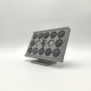 May include: A rectangular gray concrete desk clock featuring a grid of twelve circular indentations. Each indentation contains a dark gray sphere. The clock has two thin black hands centered on the face with a small gold-colored cap.
