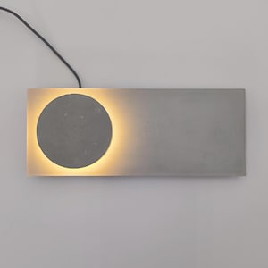 May include: A rectangular gray concrete wall light fixture featuring a circular center element. A warm, soft glow emanates from behind the circular disc, illuminating the surrounding rectangular base. A thin black power cord extends from the top edge.