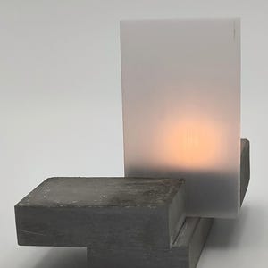 May include: A table lamp featuring a rectangular, dark gray concrete base with a smooth, matte finish. A vertical, translucent, frosted white acrylic panel is attached to the base, emitting a warm, soft orange glow from an internal light source.