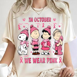 Puede incluir: Una camiseta color crema con un gráfico de Snoopy, Charlie Brown, Lucy y Linus usando gorros y bufandas rosas. El texto dice IN OCTOBER WE WEAR PINK, rodeado de lazos rosas de concienciación sobre el cáncer y pequeñas ilustraciones de corazones.