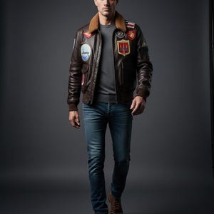 Peut inclure: Un blouson aviateur en cuir marron foncé avec un col en fausse fourrure beige, orné de multiples écussons brodés colorés sur la poitrine et les manches, porté sur un pull gris à col rond, un jean bleu et des bottines à lacets en cuir marron.