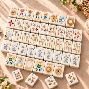 Puede incluir: Un conjunto de fichas de mahjong rectangulares blancas con ilustraciones coloridas de coronas, letras, motivos florales, corazones y símbolos numerados. Cada ficha muestra diseños detallados en oro, azul, verde y rojo.