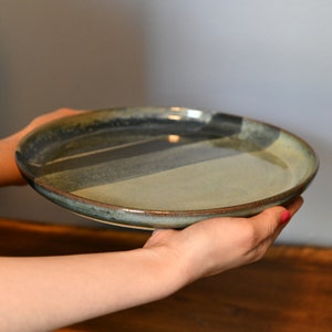 May include: A handcrafted ceramic plate with a distinctive design. The plate has a mix of blue, grey, and tan hues, with a dark brown rim. The plate is held in the hands, demonstrating its size and form.