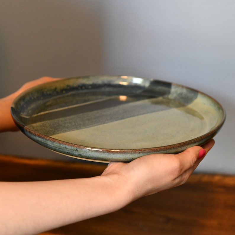May include: A handcrafted ceramic plate with a distinctive design. The plate has a mix of blue, grey, and tan hues, with a dark brown rim. The plate is held in the hands, demonstrating its size and form.
