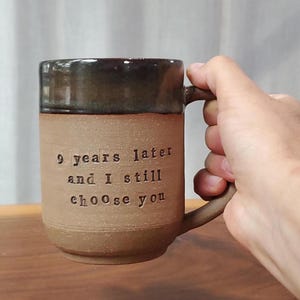 May include: A brown ceramic mug with a dark brown glazed rim and a handle. The mug features the stamped text 9 years later and I still choose you in black lettering on the front. The body of the mug has a matte, textured finish.