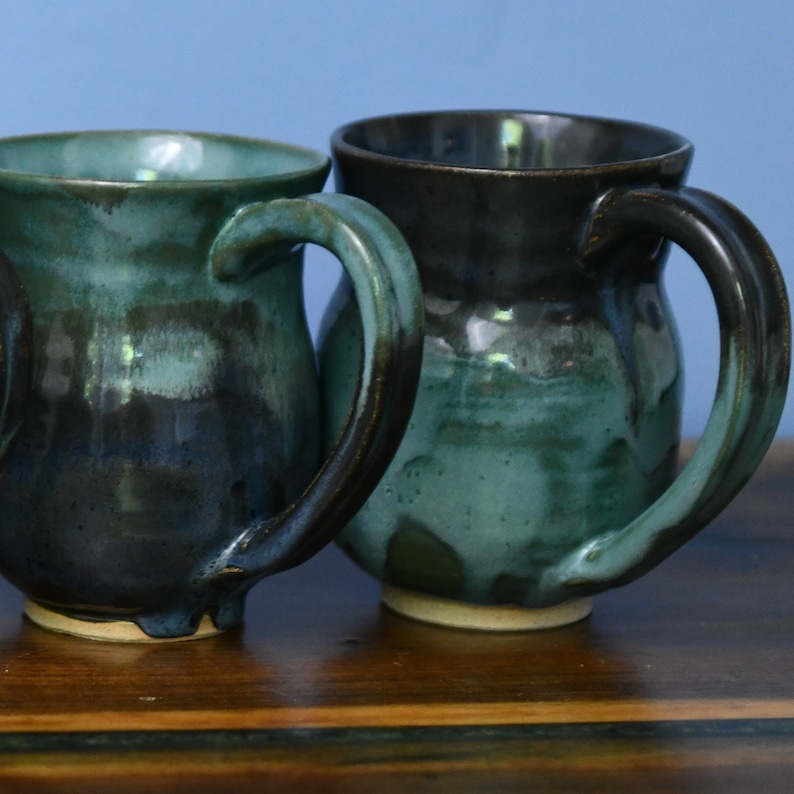 May include: Two handmade ceramic mugs with a teal and dark brown glaze. The mugs have large handles and sit on a wooden surface. The mugs are a unique addition to any kitchenware collection.