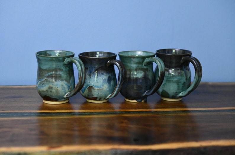 May include: Four handmade ceramic mugs with a teal and dark blue glaze. Each mug has a large handle and a unique color pattern, sitting on a dark wooden surface. The mugs are a part of a set.