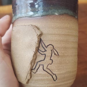 May include: A handmade ceramic mug with a textured, tan exterior and a dark brown rim. A black outline of a rock climber is etched into the side. The mug is held by a hand.