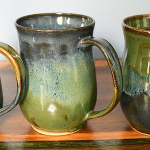 May include: Three handmade ceramic mugs with a glossy finish. The mugs feature a gradient of green, blue, and black hues. Each mug has a large handle and a wide opening, perfect for coffee or tea. The mugs are displayed on a wooden surface.