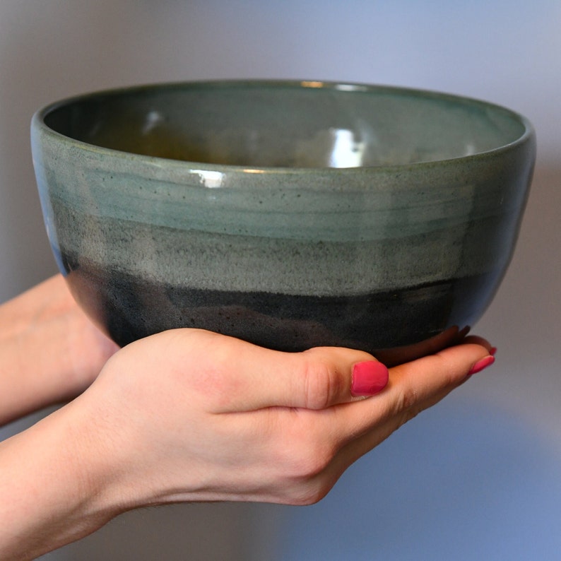 May include: A handcrafted ceramic bowl with a colour gradient, shifting from dark charcoal at the base to teal green at the rim. The bowl is held in two hands, highlighting its smooth, rounded form. Suitable for serving or as a decorative accent.