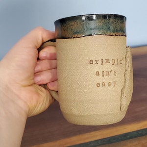 Puede incluir: Una taza de cerámica hecha a mano con un cuerpo beige y un borde verde oscuro y negro. La taza presenta el texto "crimpin' ain't easy" en una fuente estampada. Un elemento decorativo texturizado está en el lateral de la taza.