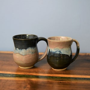 May include: Two handmade ceramic mugs with a unique glaze. One mug is dark with a blue and tan band, the other is brown with a blue and tan band. Both mugs have a curved handle and sit on a wooden surface.